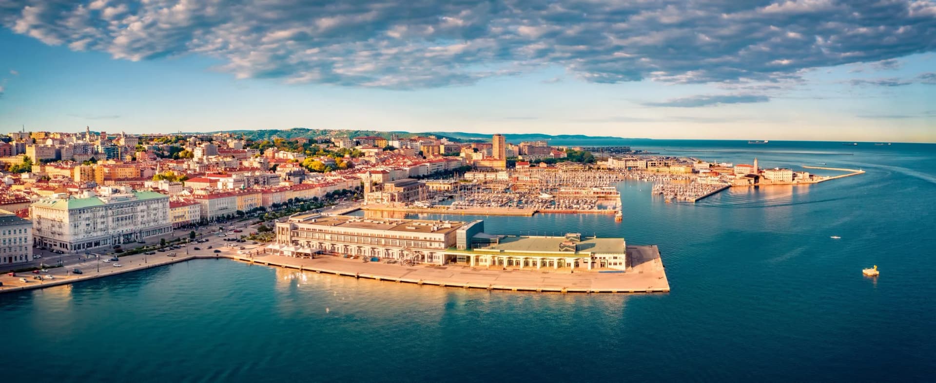 Aerial view of Trieste city waterfront, harbor filled with boats, and blue sea under cloudy sky.