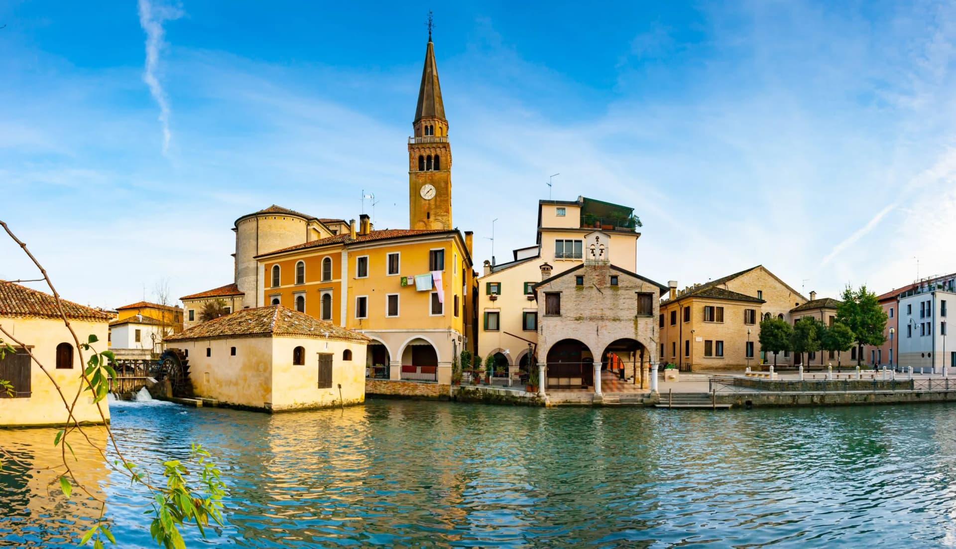 Historic buildings along a canal with a tall clock tower in Portogruaro under a blue sky.