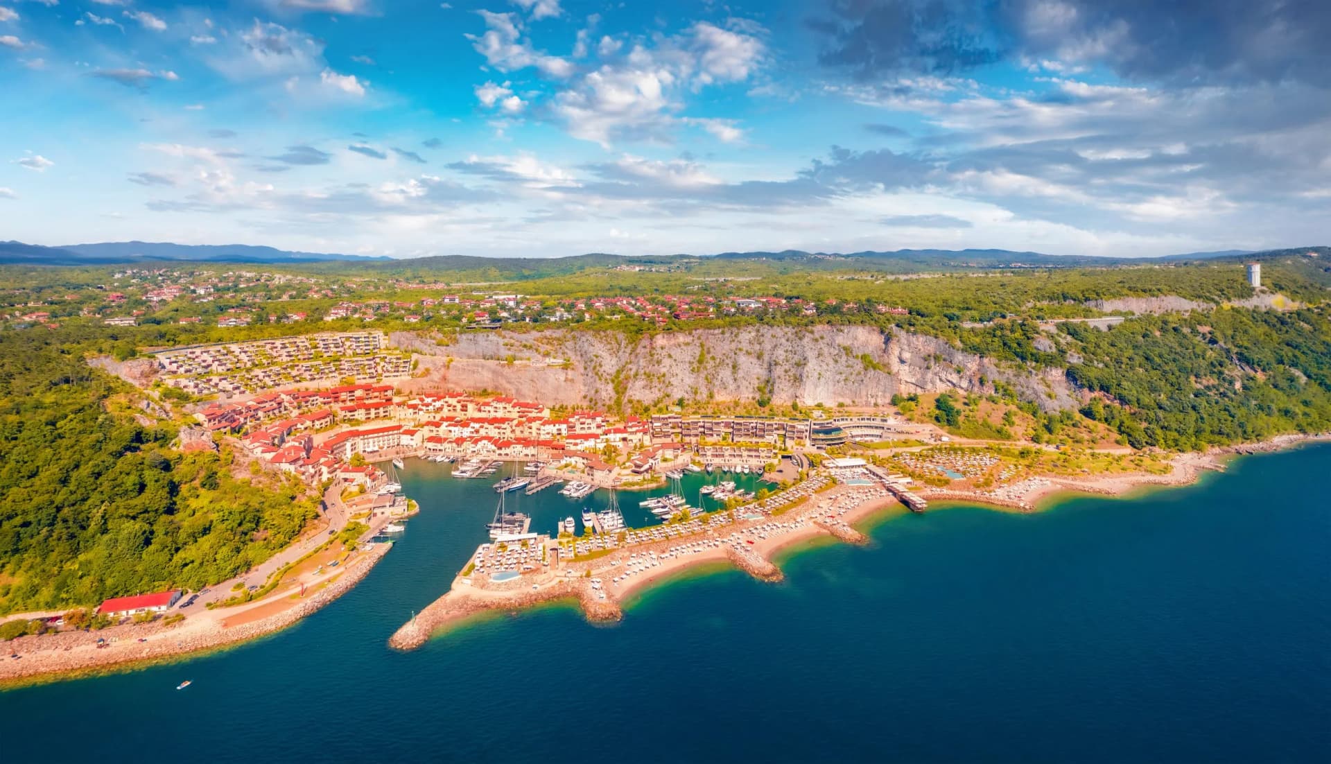 Coastal resort town with red-roofed buildings, marina, and beach below a steep, green hillside.