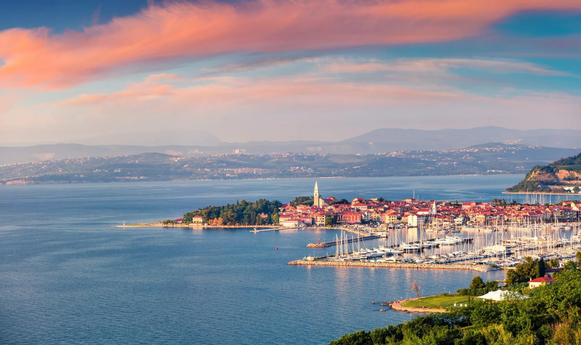 Coastal town with red roofs and marina full of sailboats at sunset near mountains