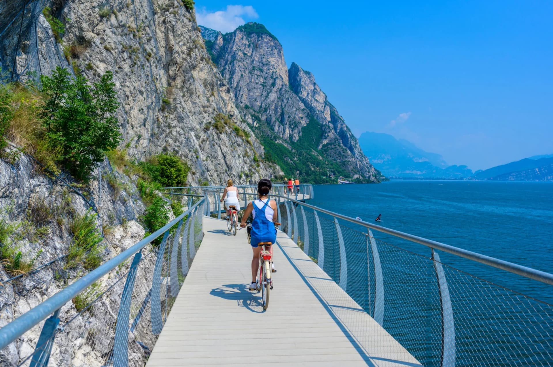 Cyclists on a boardwalk path beside steep mountains overlooking a large blue lake, Lombardy.