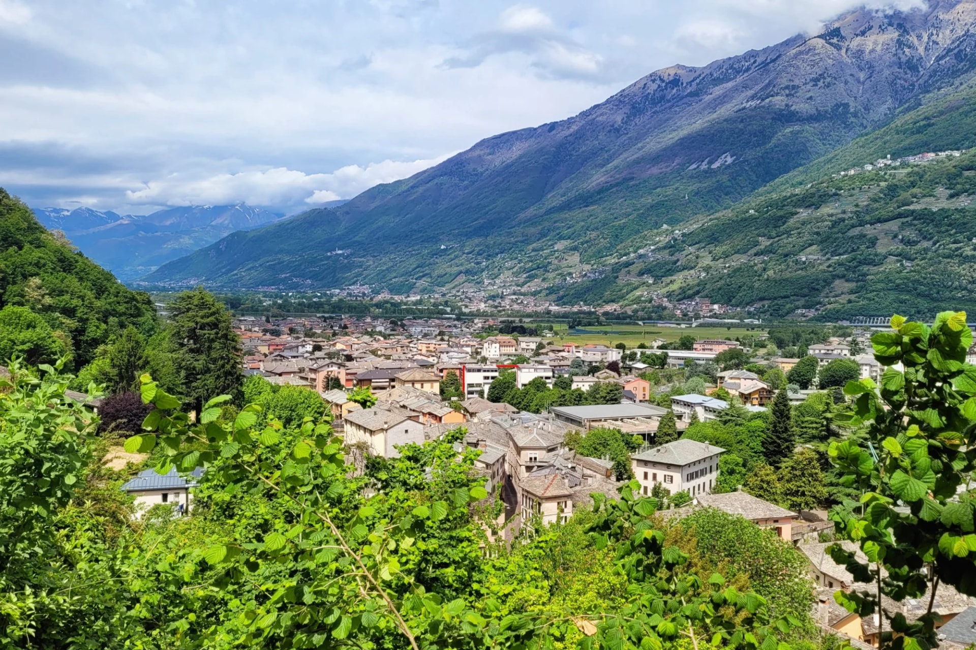 Town of Morbegno in Valtellina valley with steep green mountains under cloudy sky.