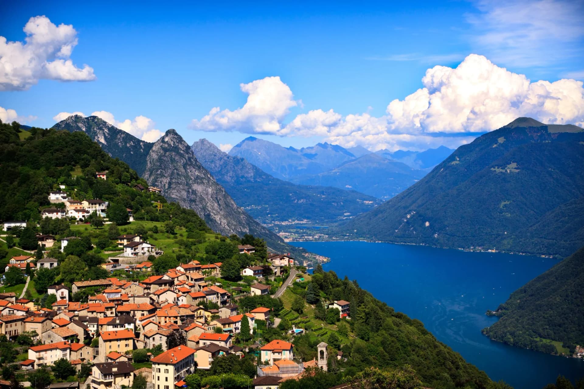 Village on steep green hillside overlooking deep blue Lake Lugano and surrounding Alps.