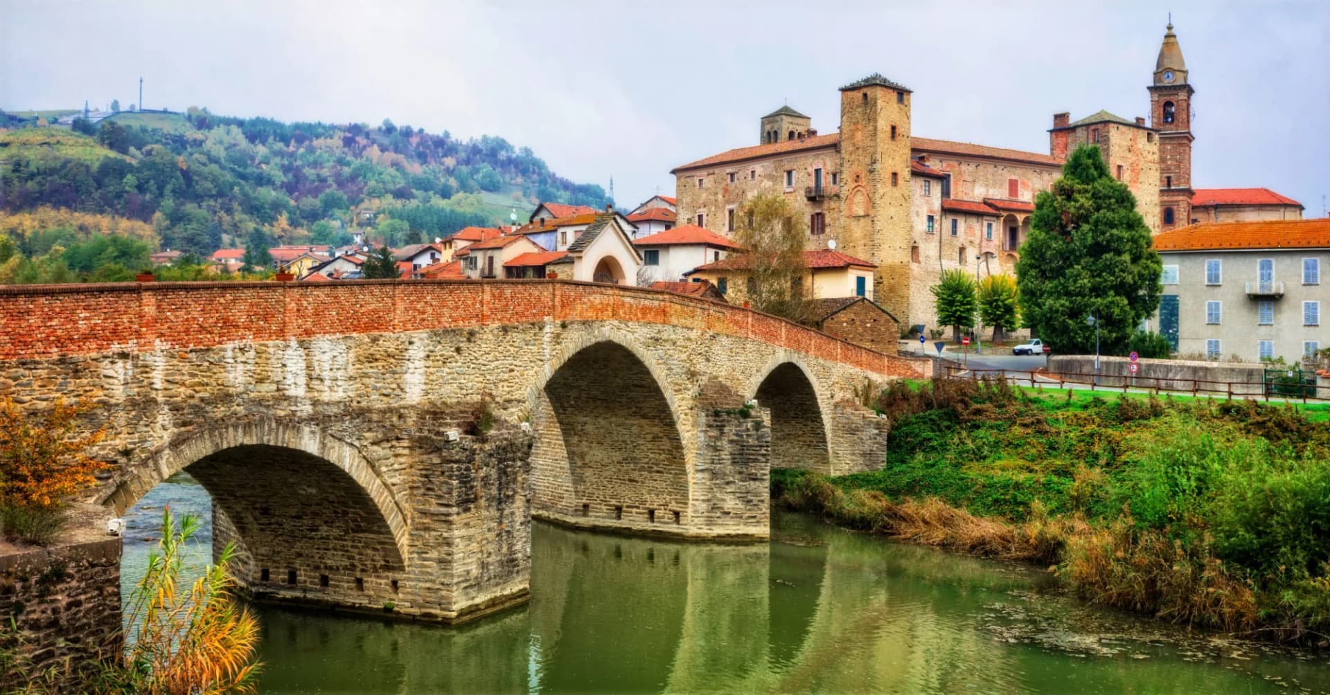 Stone arch bridge over green river leading to historic buildings in Regione Asti, Piemonte, Italy.