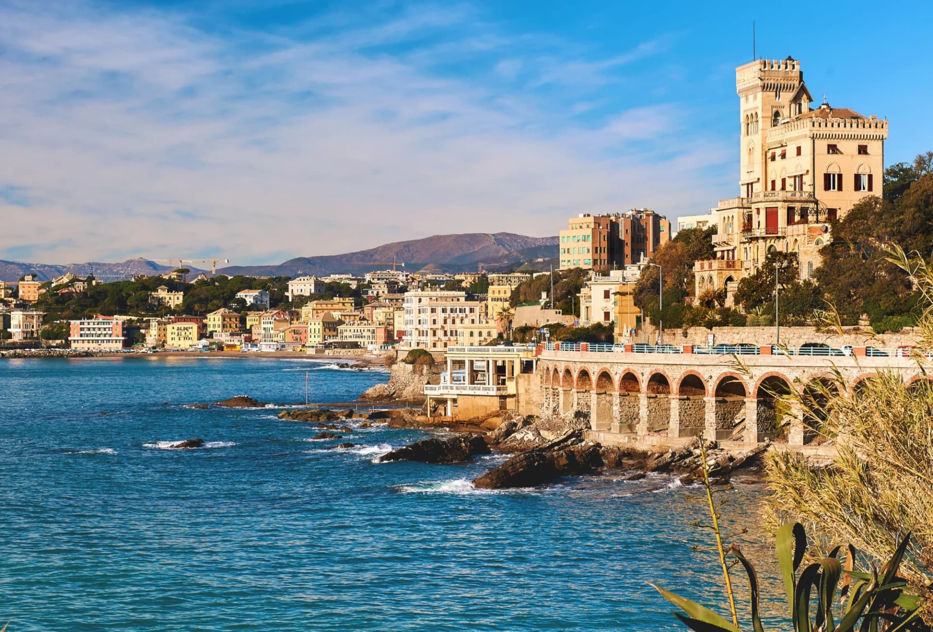 Coastal view of Genoa with colorful buildings, a castle-like tower, and mountains in the background.