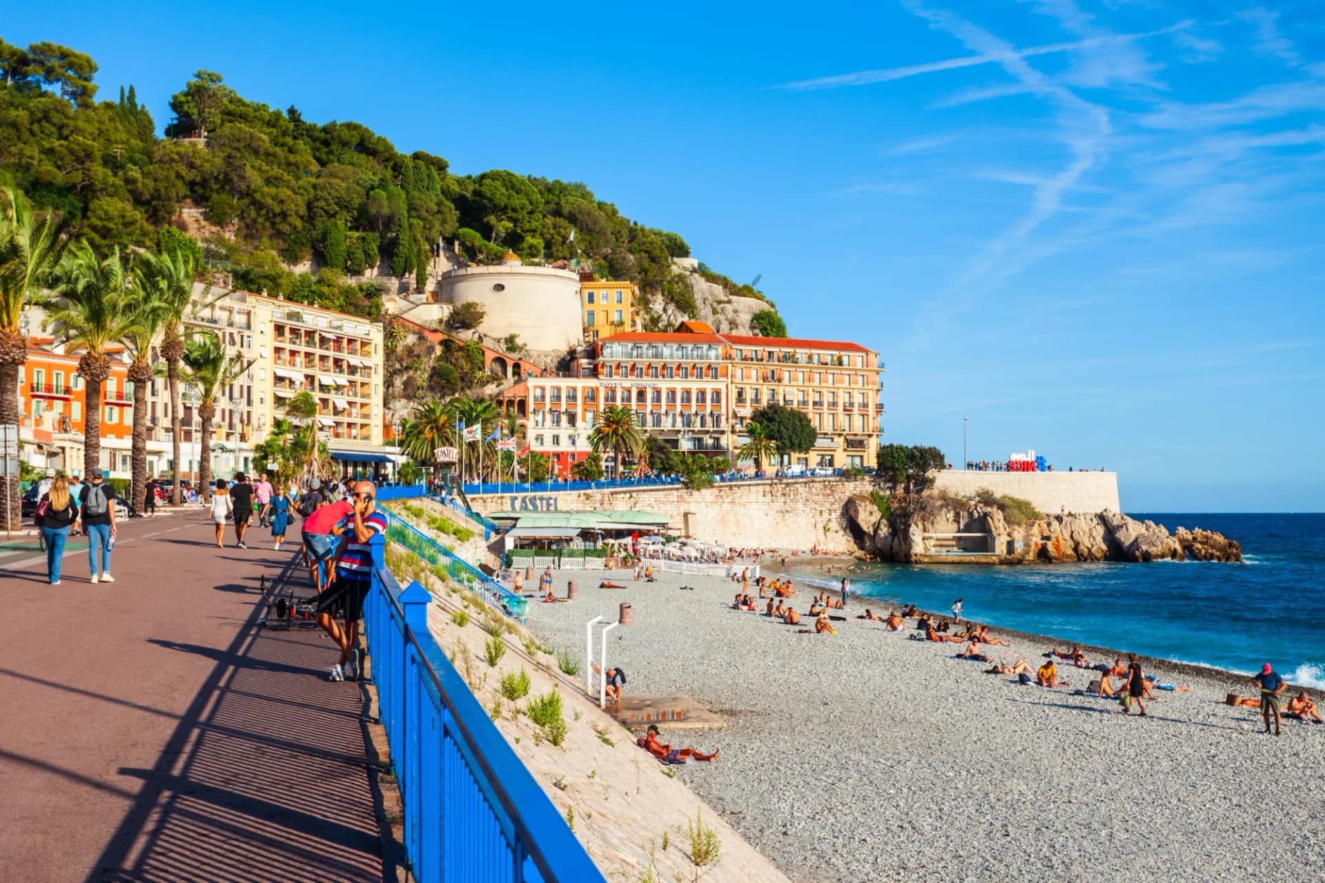 Promenade and pebble beach with sunbathers below a green hillside in Nice, France.