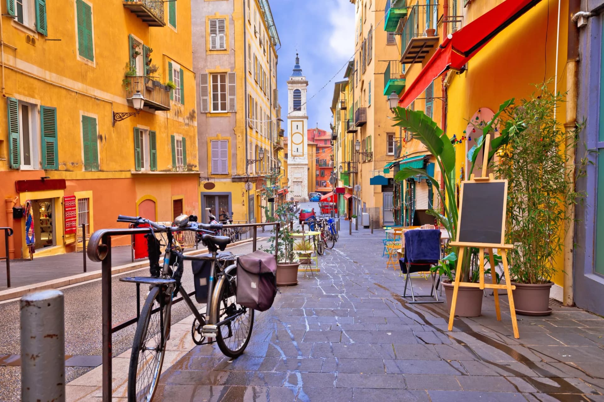 Colorful street architecture leading to a church tower with a bicycle parked on wet cobblestones.
