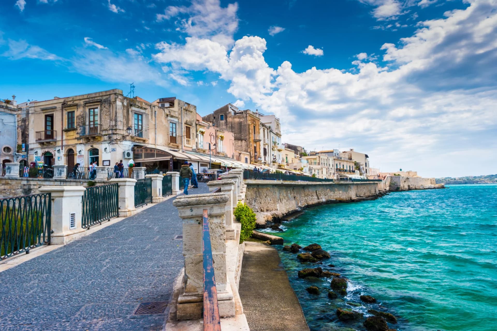 Cobblestone walkway along turquoise sea next to historic buildings on the coast of Ortigia Island, Syracuse, Sicily.