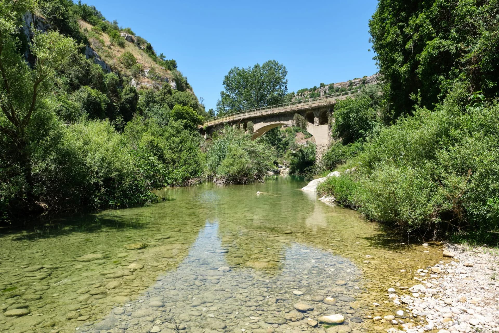 River with clear, shallow water and rocky bottom under an arched bridge in Parco Naturale di Pantalica.