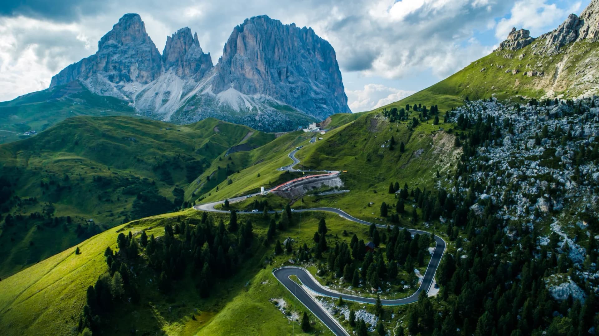 Winding mountain road through green slopes toward jagged peaks in the Dolomites.