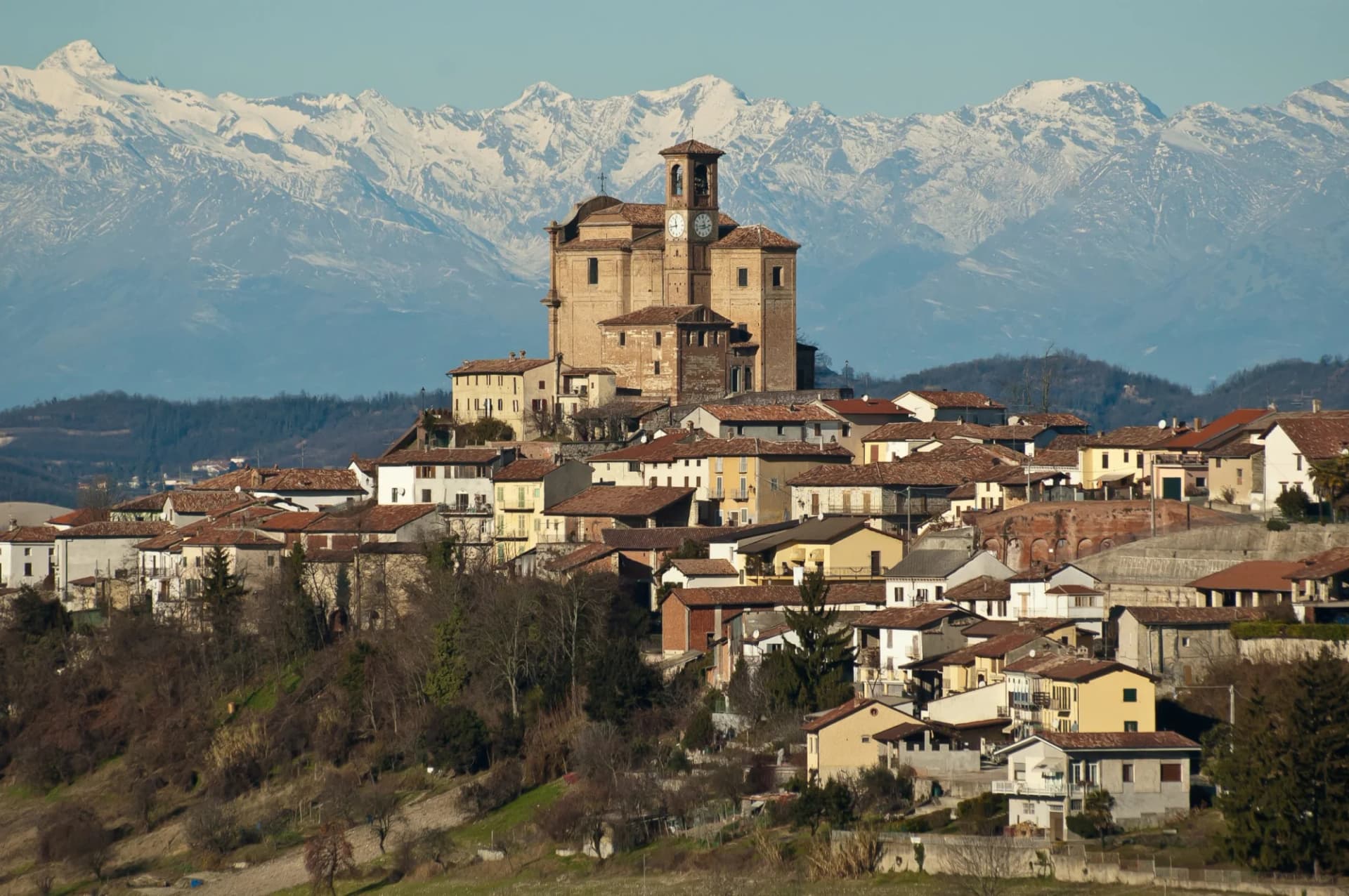 Hilltop village with terracotta roofs below snow-capped mountains, Monferrato region.