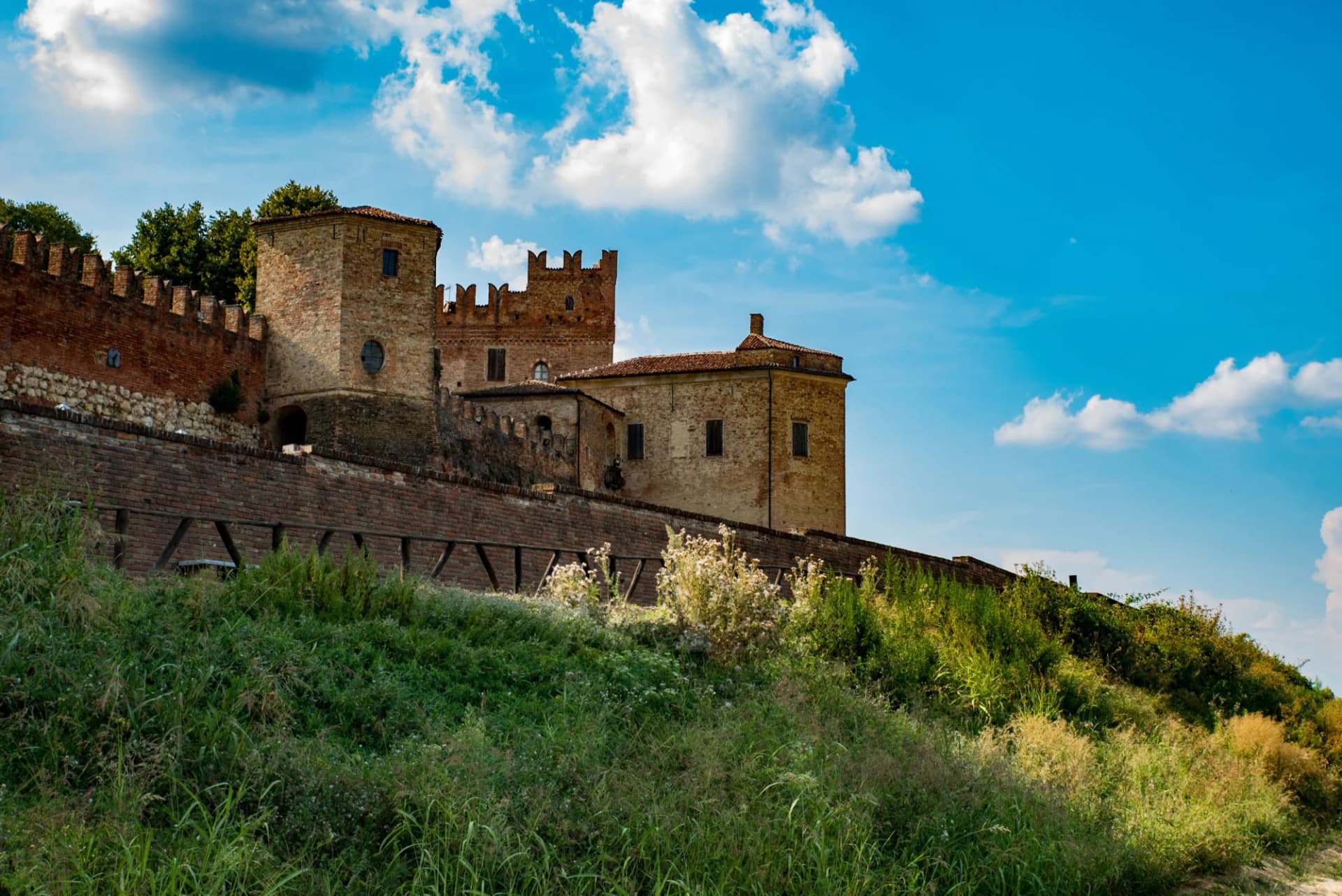 Brick castle ruins with crenelated walls rising above overgrown grassy embankment under blue sky.