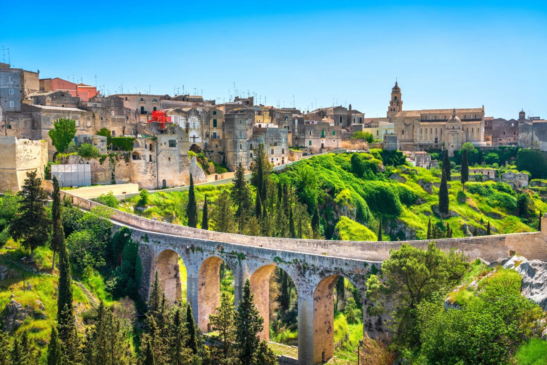 Stone bridge over a canyon leading to the ancient town of Gravina in Puglia with lush green hills.