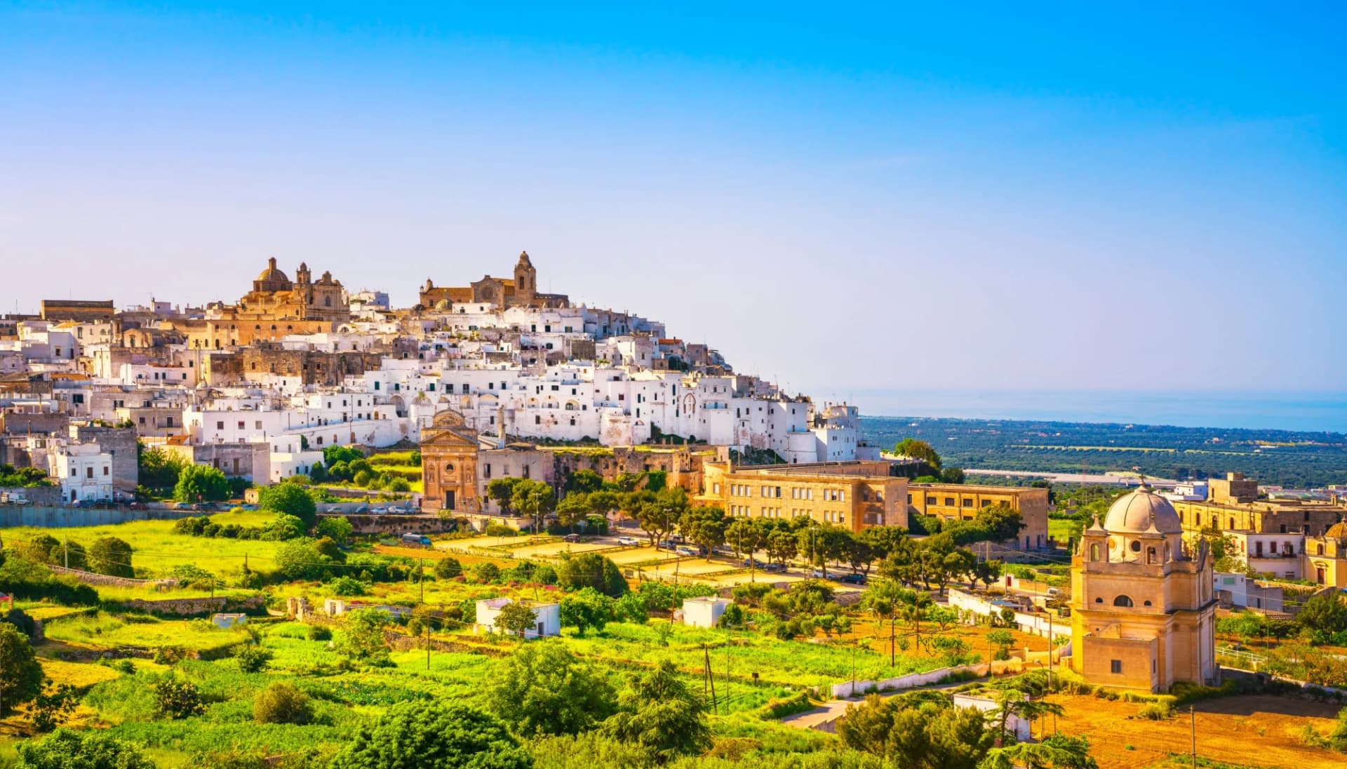 Ostuni white town skyline and historic church overlooking green fields and the sea in Brindisi, Apulia, Italy.