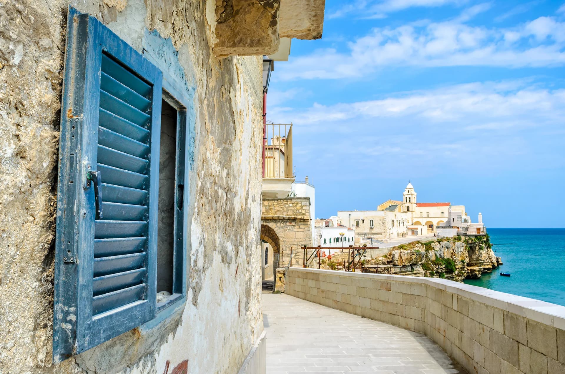 Stone pathway in Vieste, Apulia, with blue shutter and church overlooking the Mediterranean Sea.