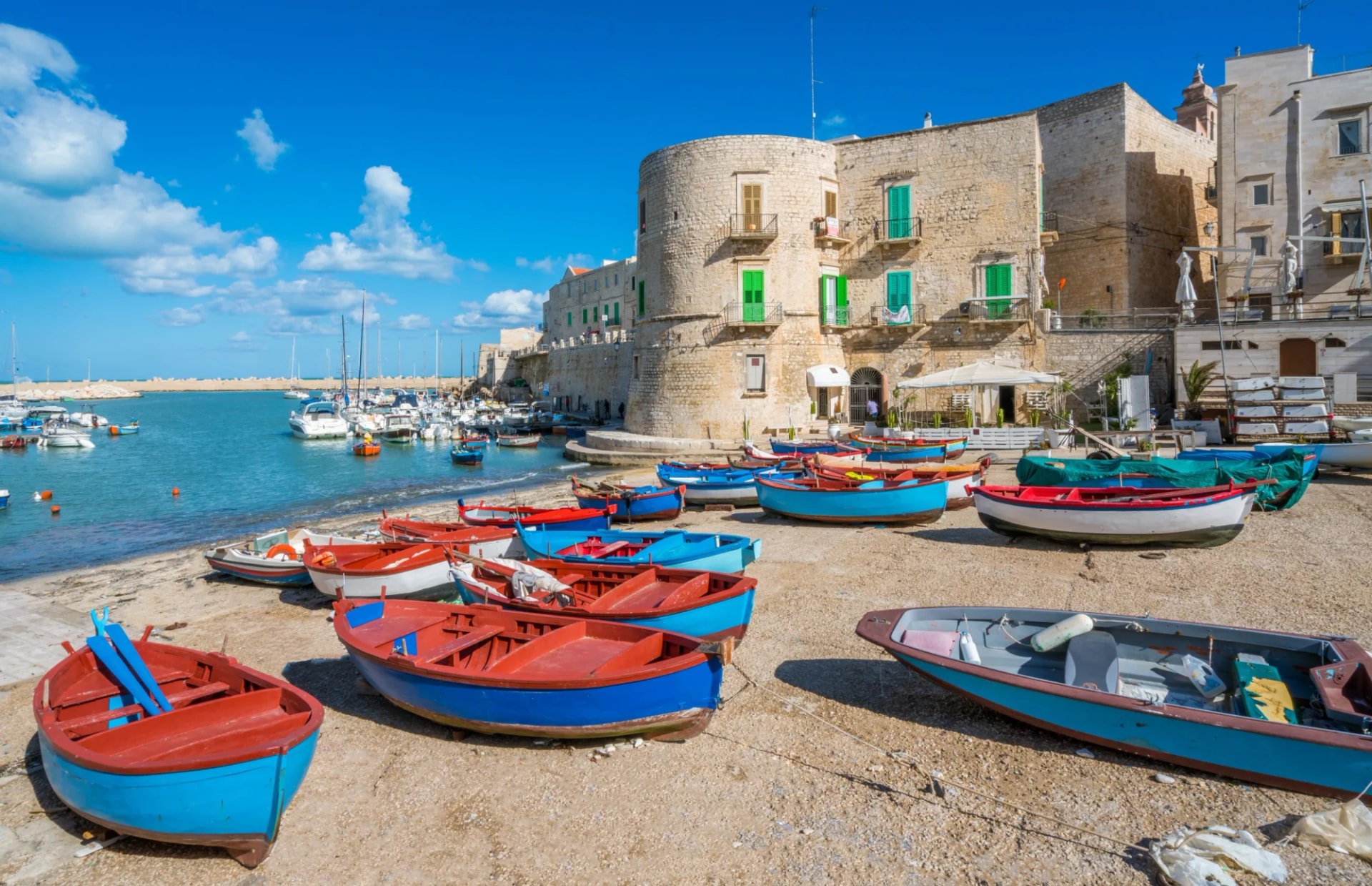 Colorful fishing boats pulled onto shore near historic stone buildings in Giovinazzo harbour, Puglia.