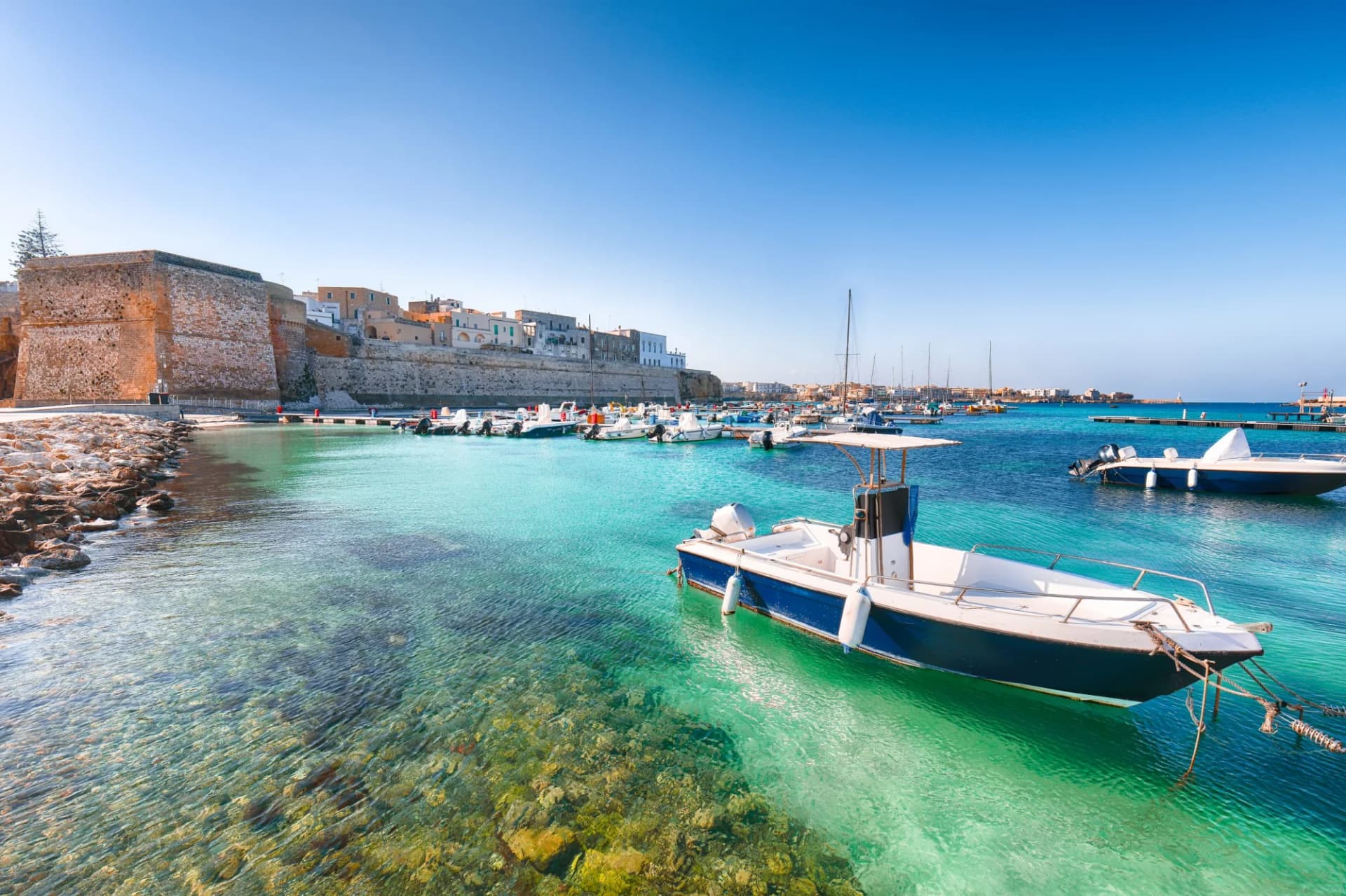 Fishing boats in turquoise harbor water near Otranto coastal town walls and buildings.