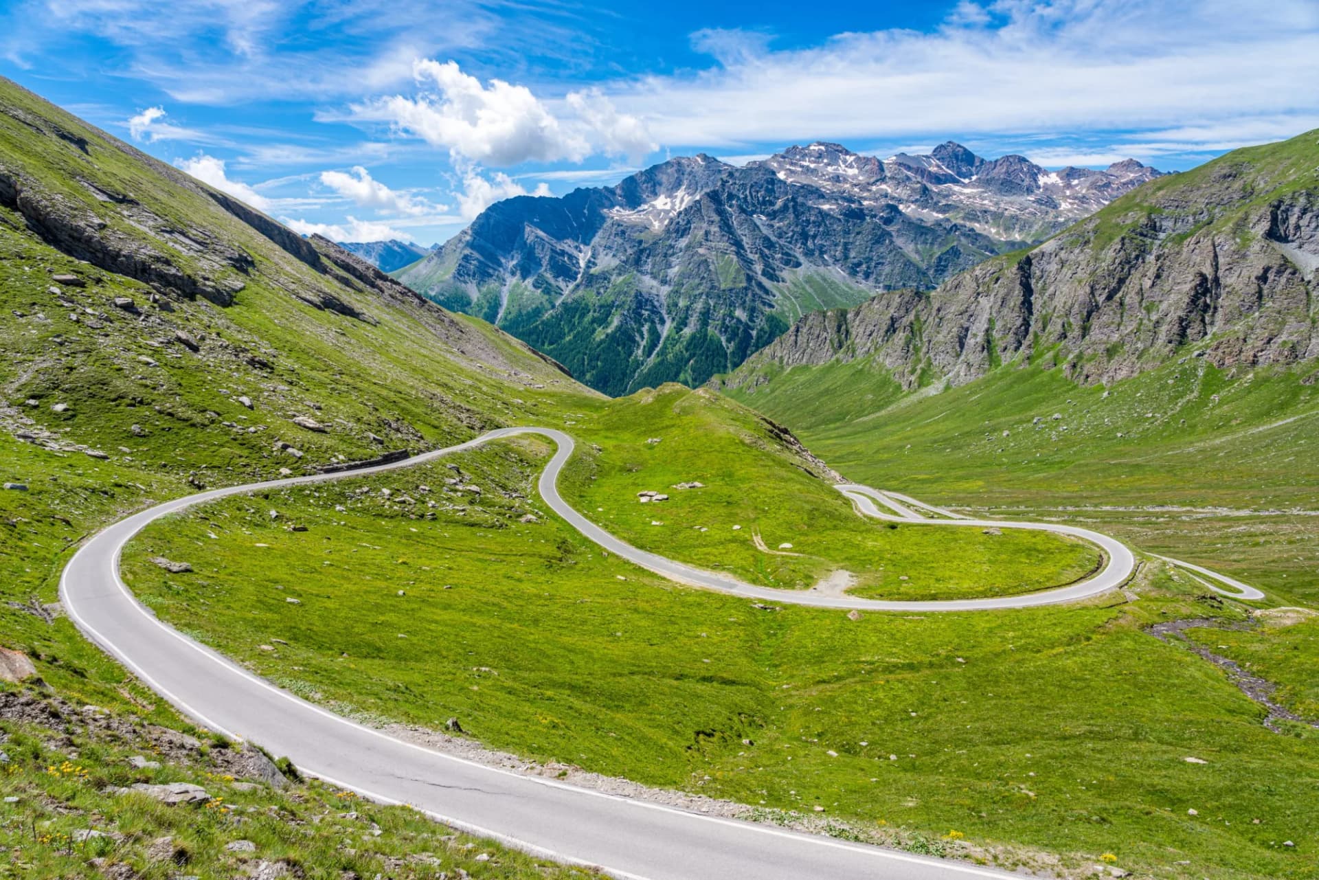 Winding mountain road ascending through green alpine slopes toward snow-capped peaks under blue sky.