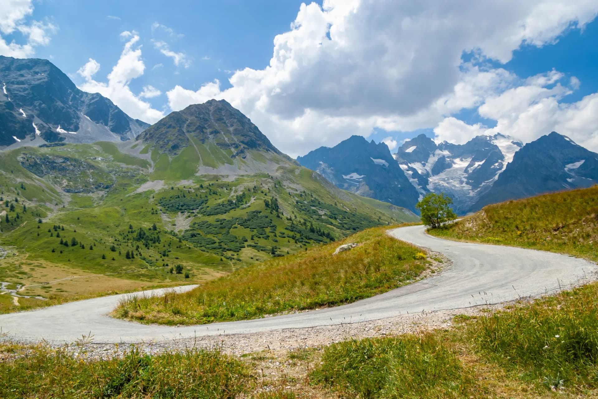Winding mountain road curve through green alpine meadows toward snow-capped peaks under a blue sky.