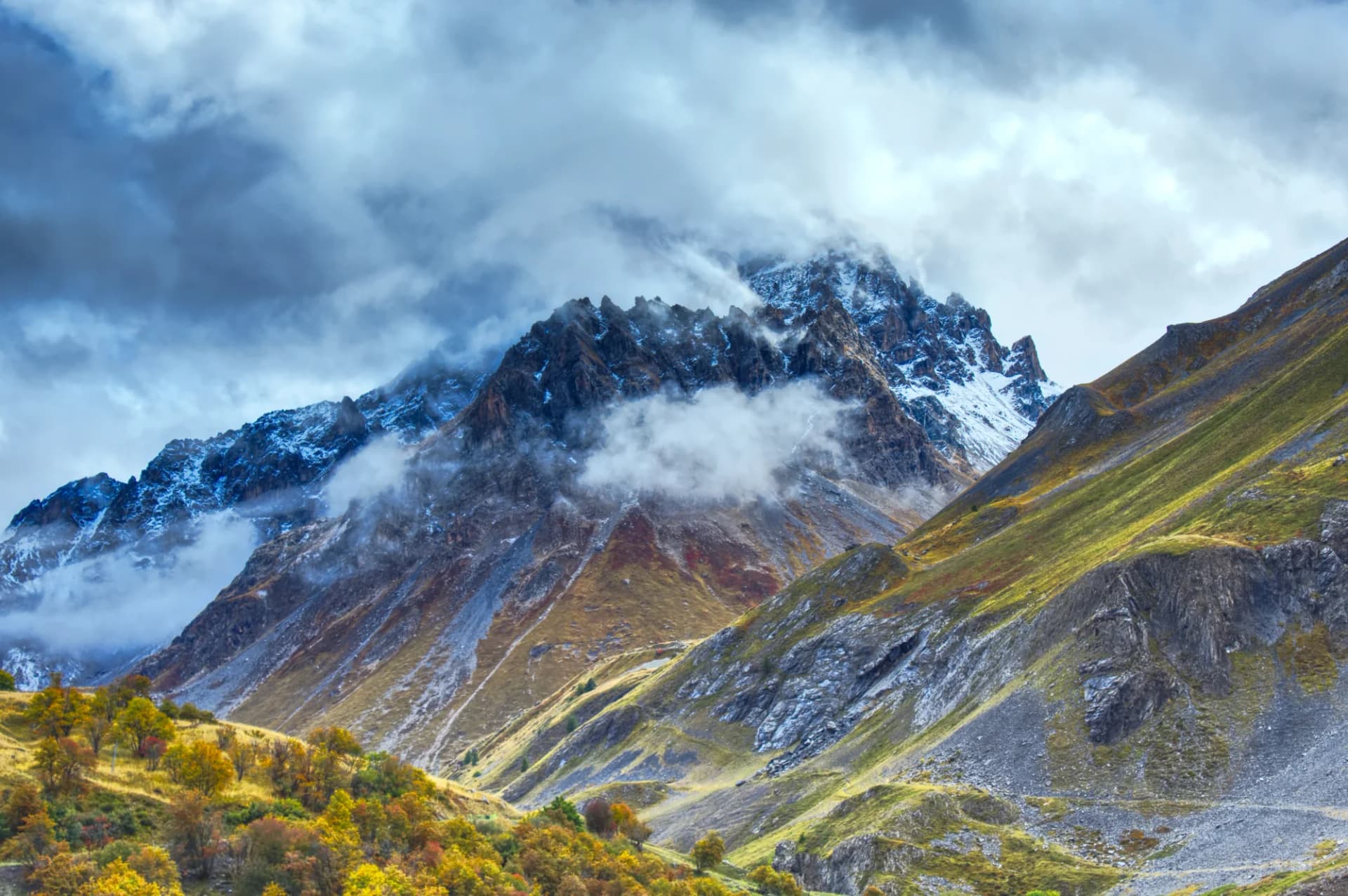 Large mountain peak with snow and clouds, surrounded by autumn foliage in the valley.