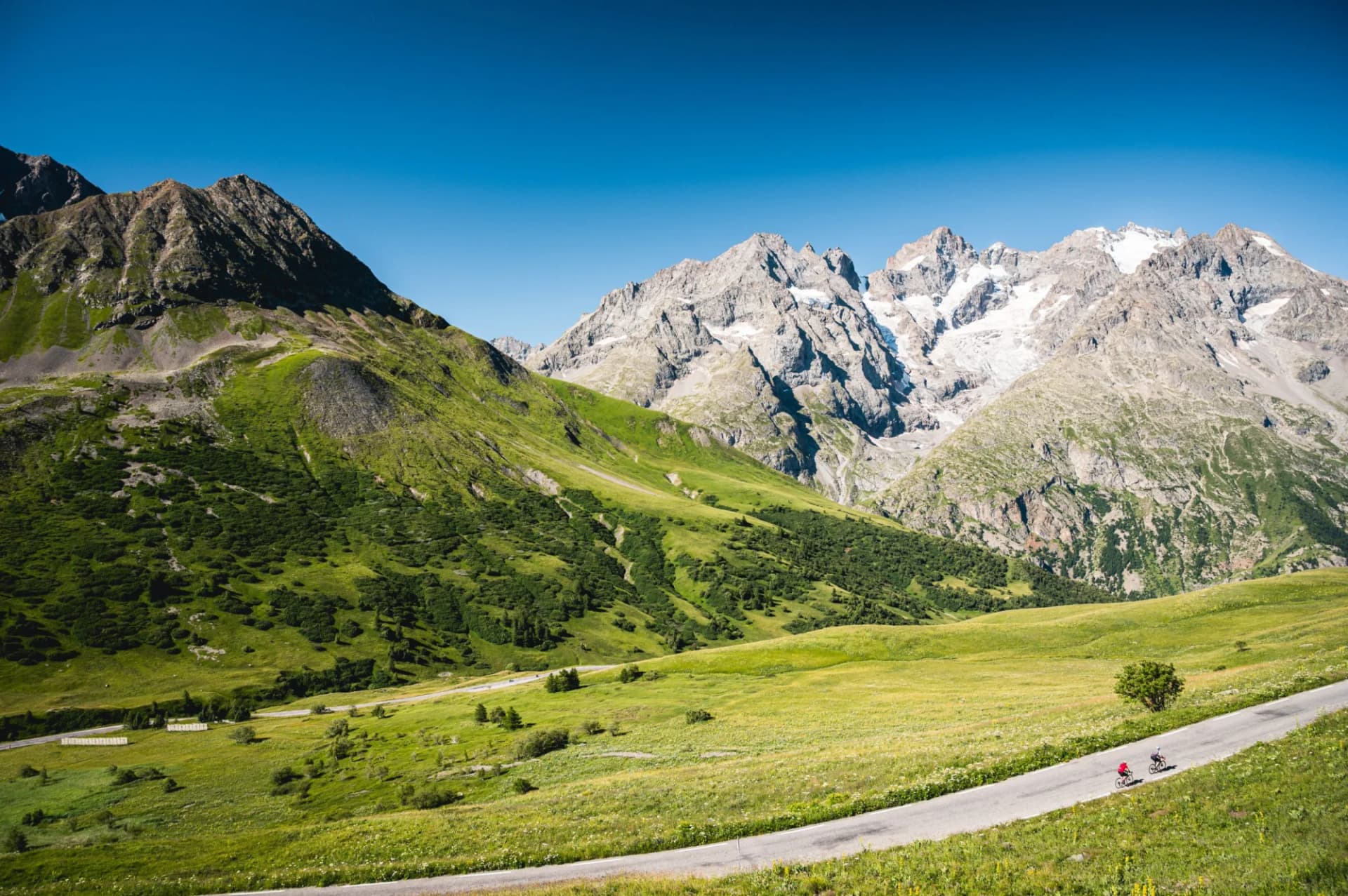 Cyclists ascending road past green slopes toward snow-capped mountains near Col du Lautaret, Hautes-Alpes.