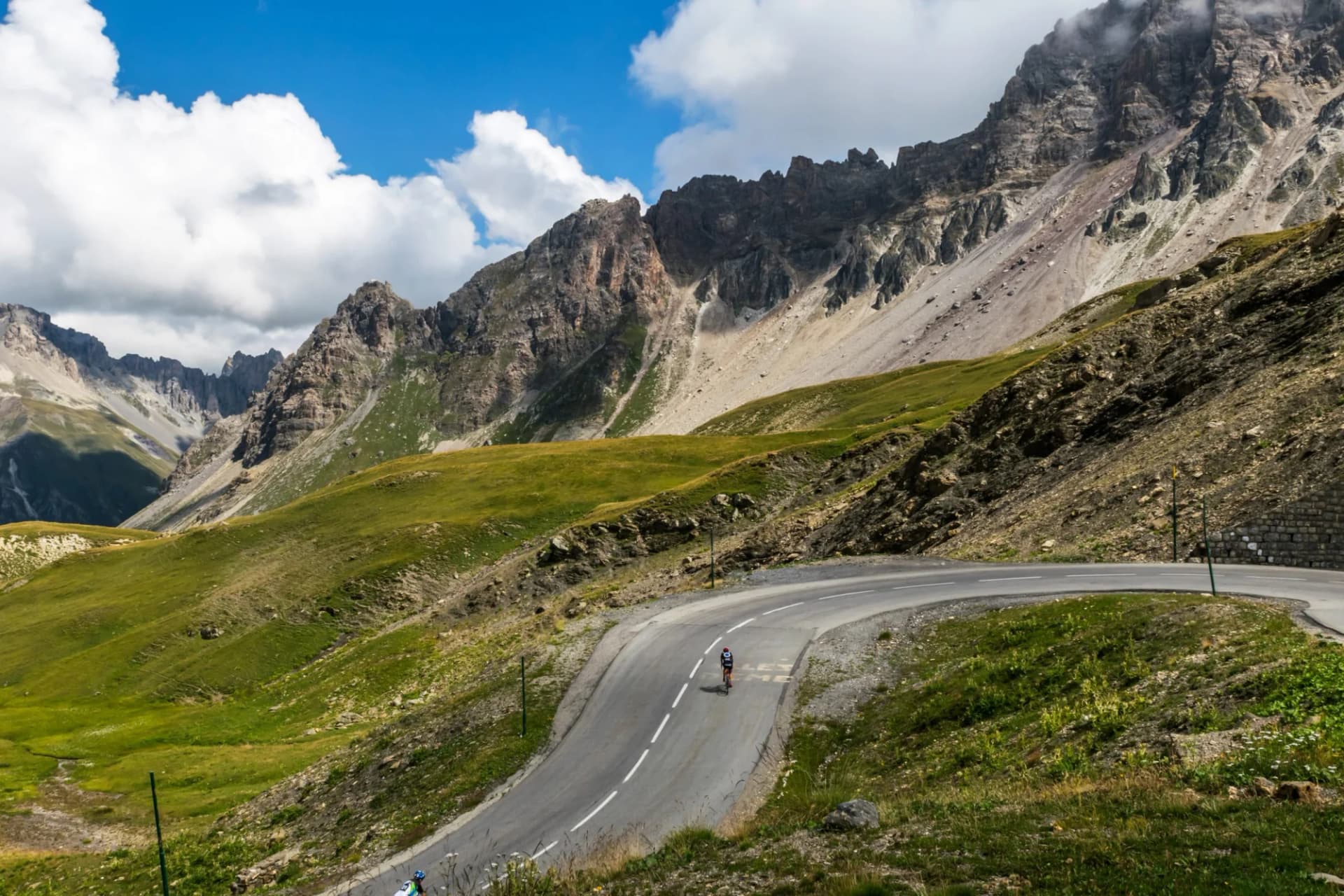 Cyclist climbing winding mountain road with steep green slopes and rocky peaks, Col du Galibier.