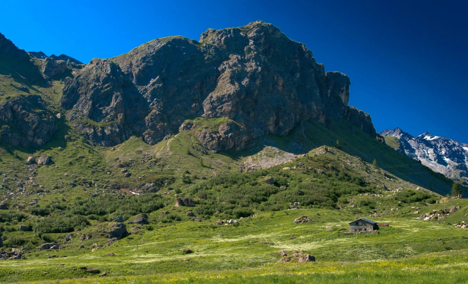 Stone hut in green alpine meadow below massive rocky mountain in Cormet de Roselend, Savoie, France.