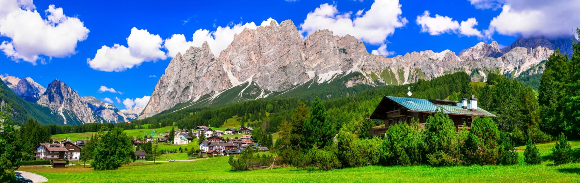 Alpine village nestled in green valley below rugged Dolomite mountains under blue sky