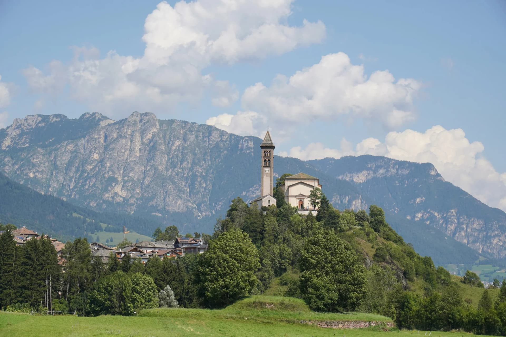 Church with bell tower on a green hill above Predazzo village and steep mountains.