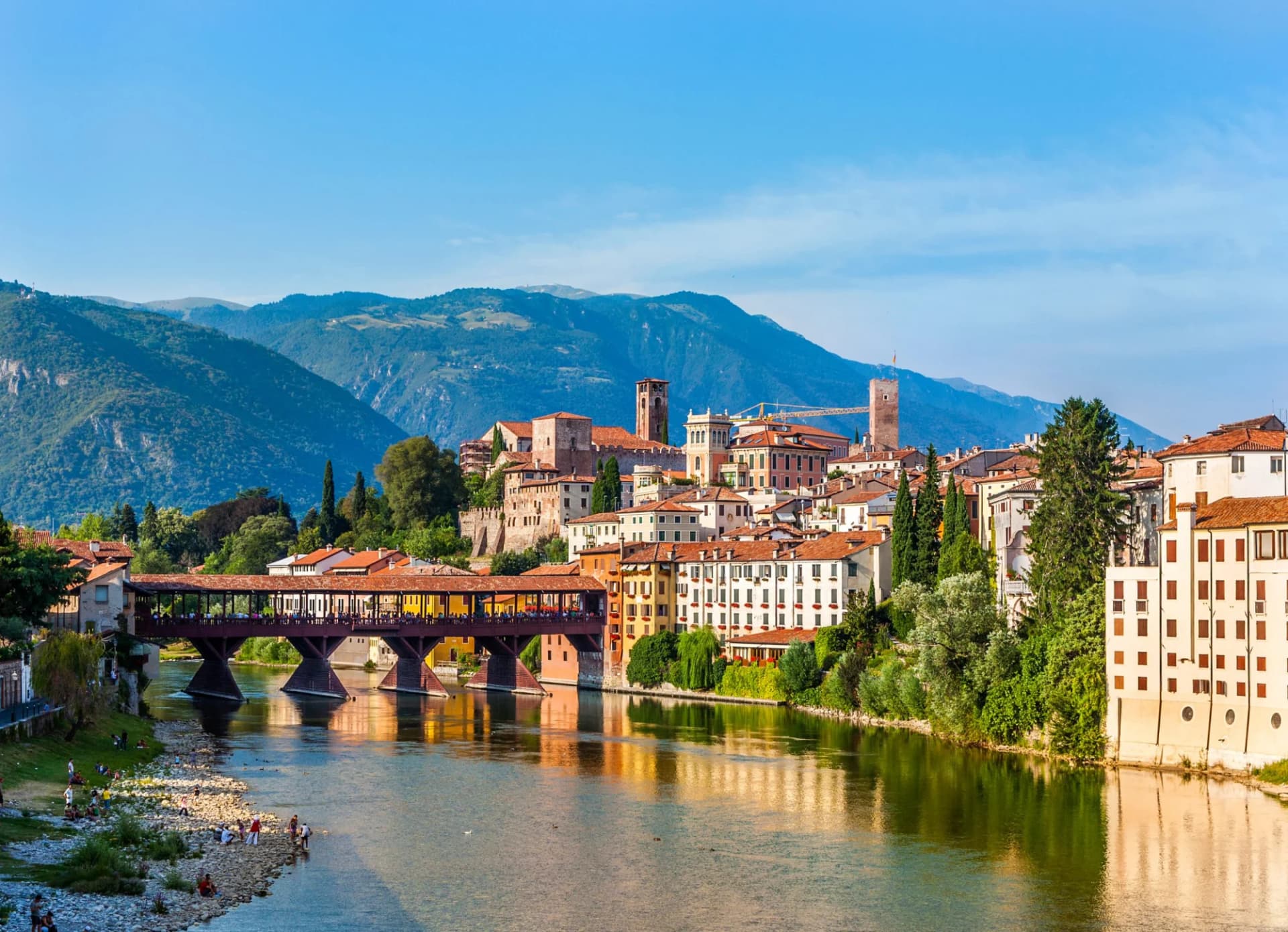 Covered wooden bridge over river in Bassano del Grappa with town and mountains backdrop.