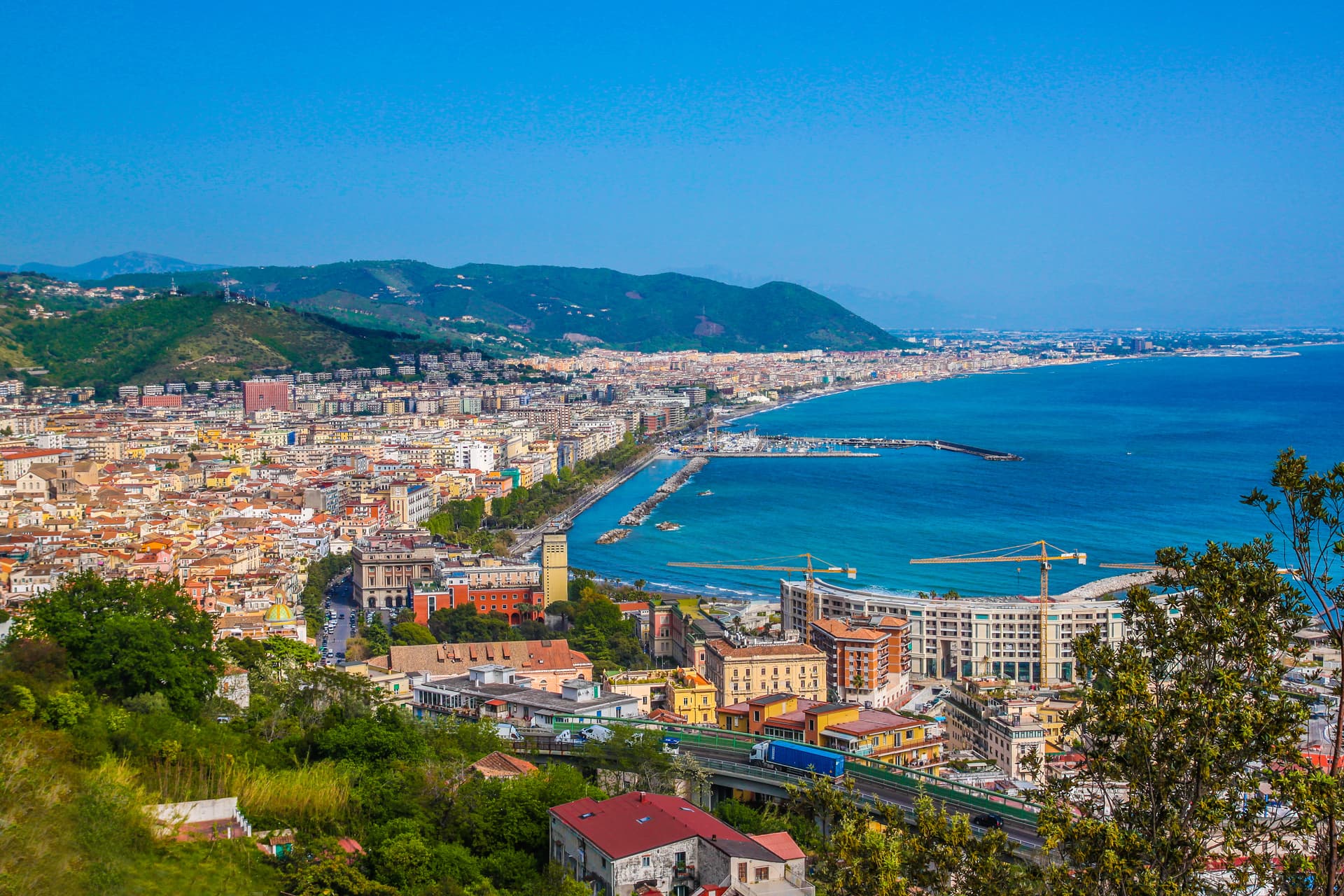 Coastal city of Salerno with dense buildings, harbor, and green mountains under a clear blue sky.