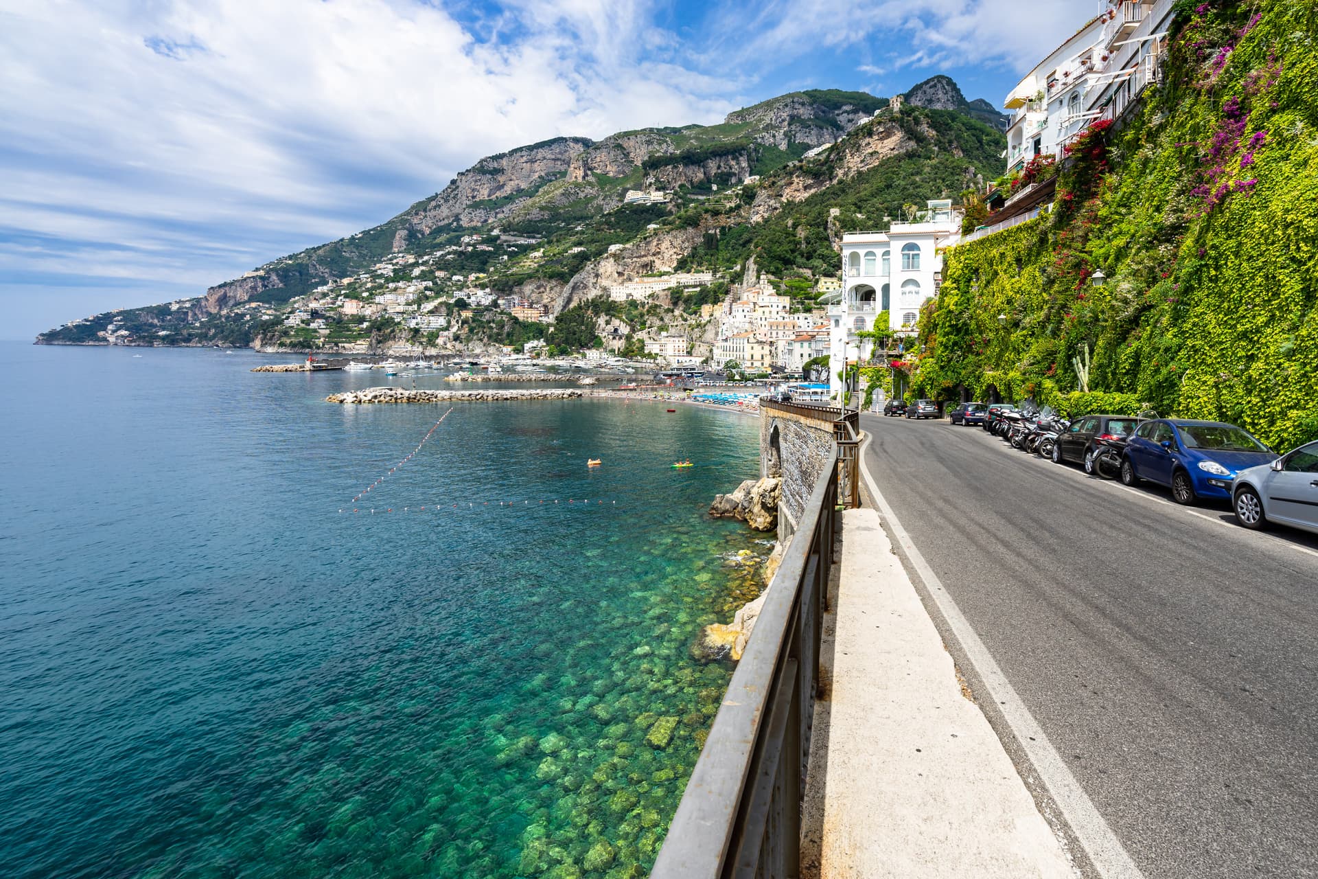 Coastal road with parked cars overlooking clear blue water and colorful buildings on a steep hillside.