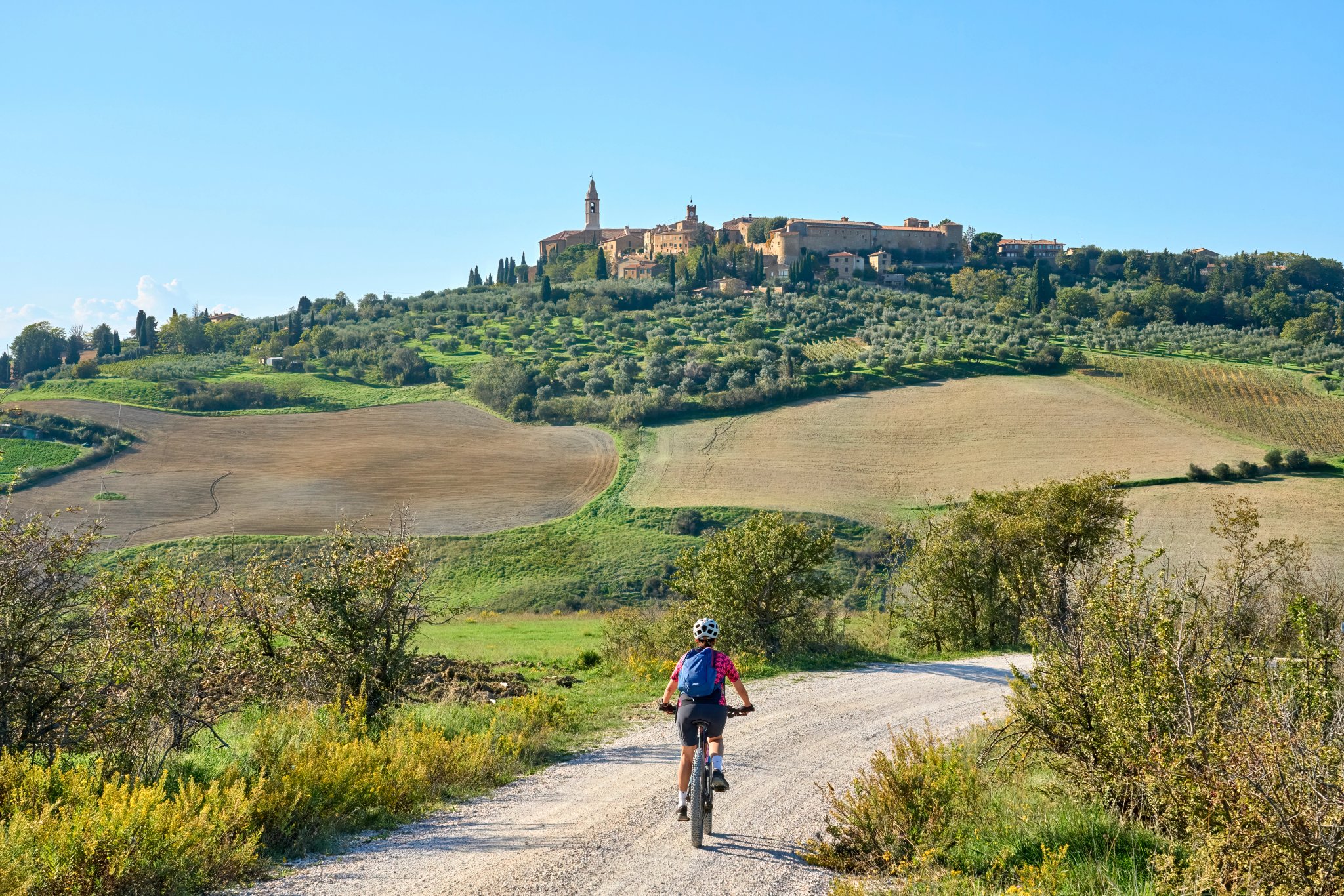 fin senior kvinde cykler på sin elektriske mountainbike i Chianti-området under skyline af den middelalderlige by Pienza, Toscana, Italien