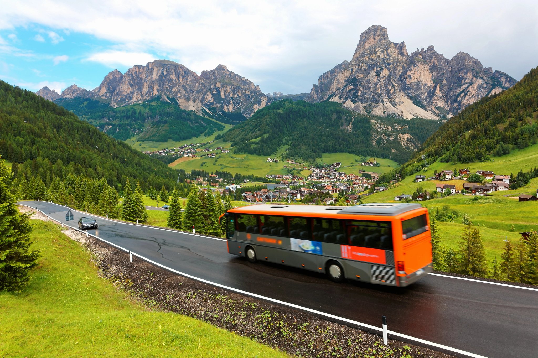 Cars & buses traveling on a scenic highway thru a green grassy valley with villages on the hillside of rugged mountains after a summer shower in Corvara, Badia, Dolomites, Trentino, South Tyrol, Italy